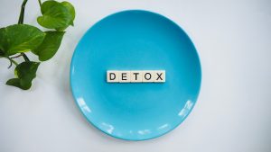 The word detox spelled with wooden letters on a blue plate next to green leaves.