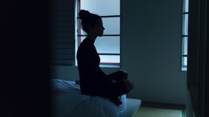 A woman sitting on a bed in a dark room with windows behind her.