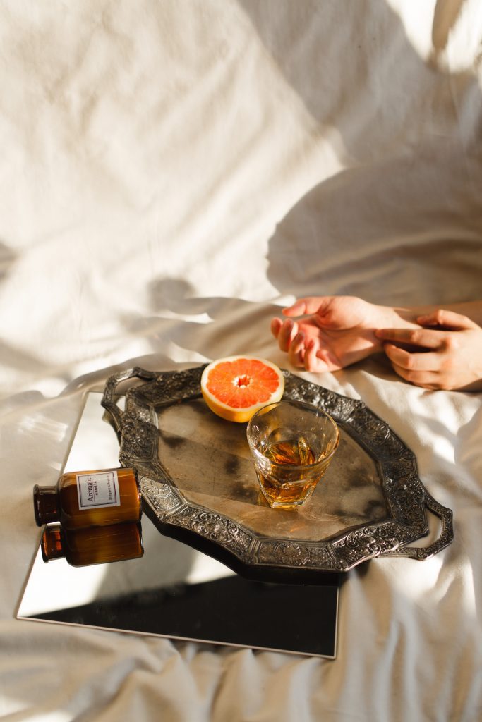 An alcoholic drink in a glass next to a half orange put on a tray that's on a mirror with an empty medicine bottle, all on bed sheets next to two hands.