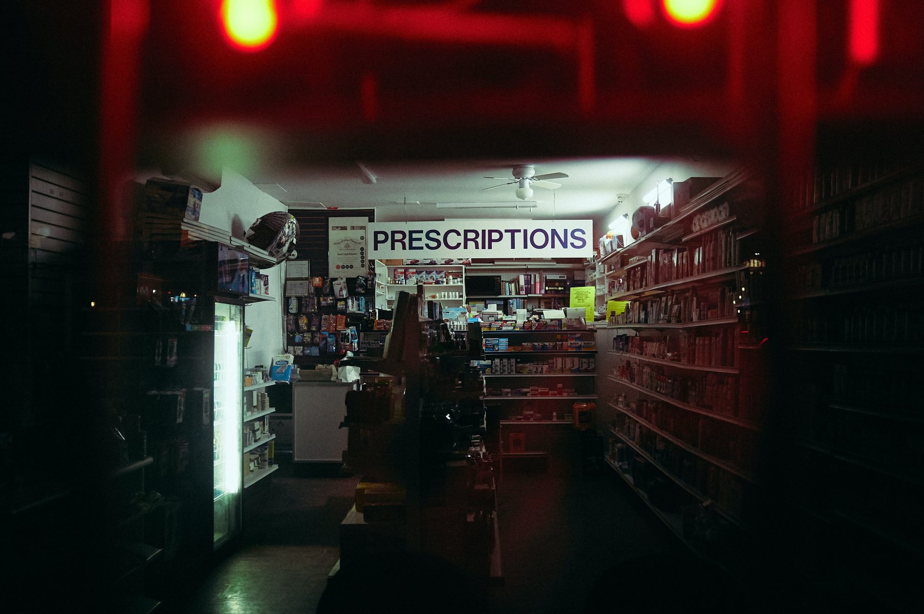 Dark picture of a small shop with various items on the shelves and a sign that spells prescriptions in the middle.