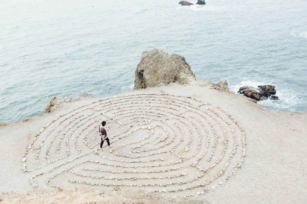 A labyrinth drawn on sand with a person walking in the middle next to the sea.