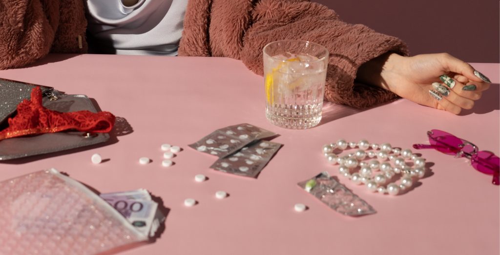 Drink, pills, purse with money sticking out on a pink table with a hand resting next to them.